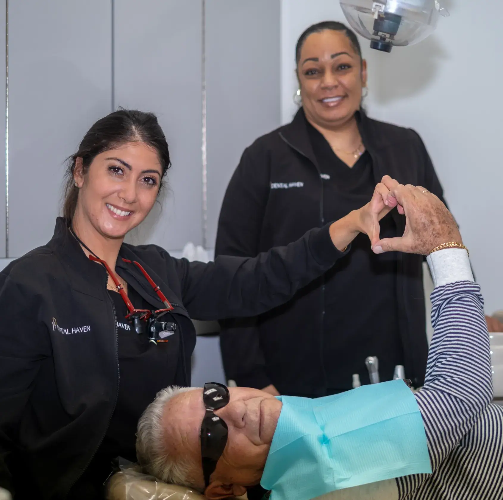 Two dental professionals smile as a patient makes a heart shape with hands in the chair.