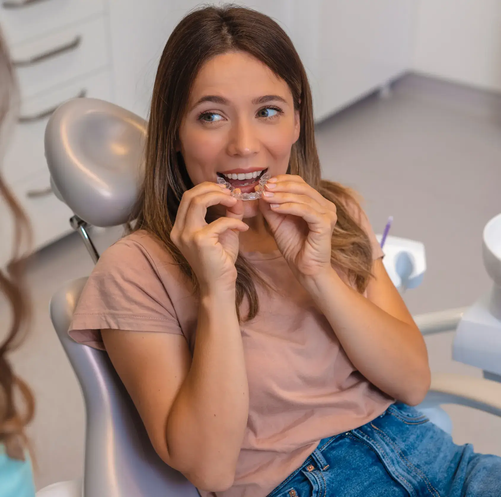 A woman sitting in a dentist's chair is fitting a clear dental aligner onto her teeth.