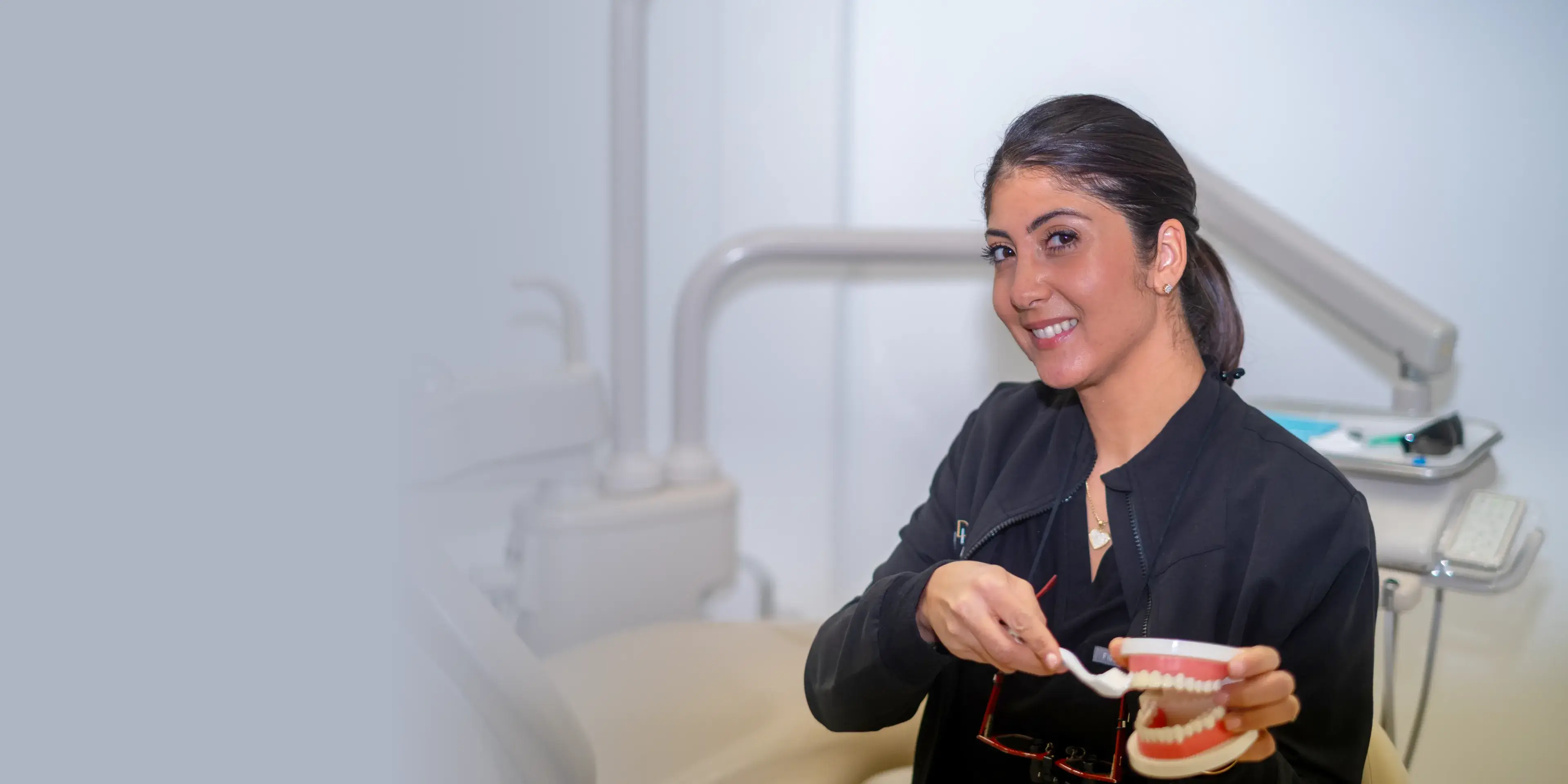 A woman smiles while demonstrating tooth brushing on a dental model in an office setting.