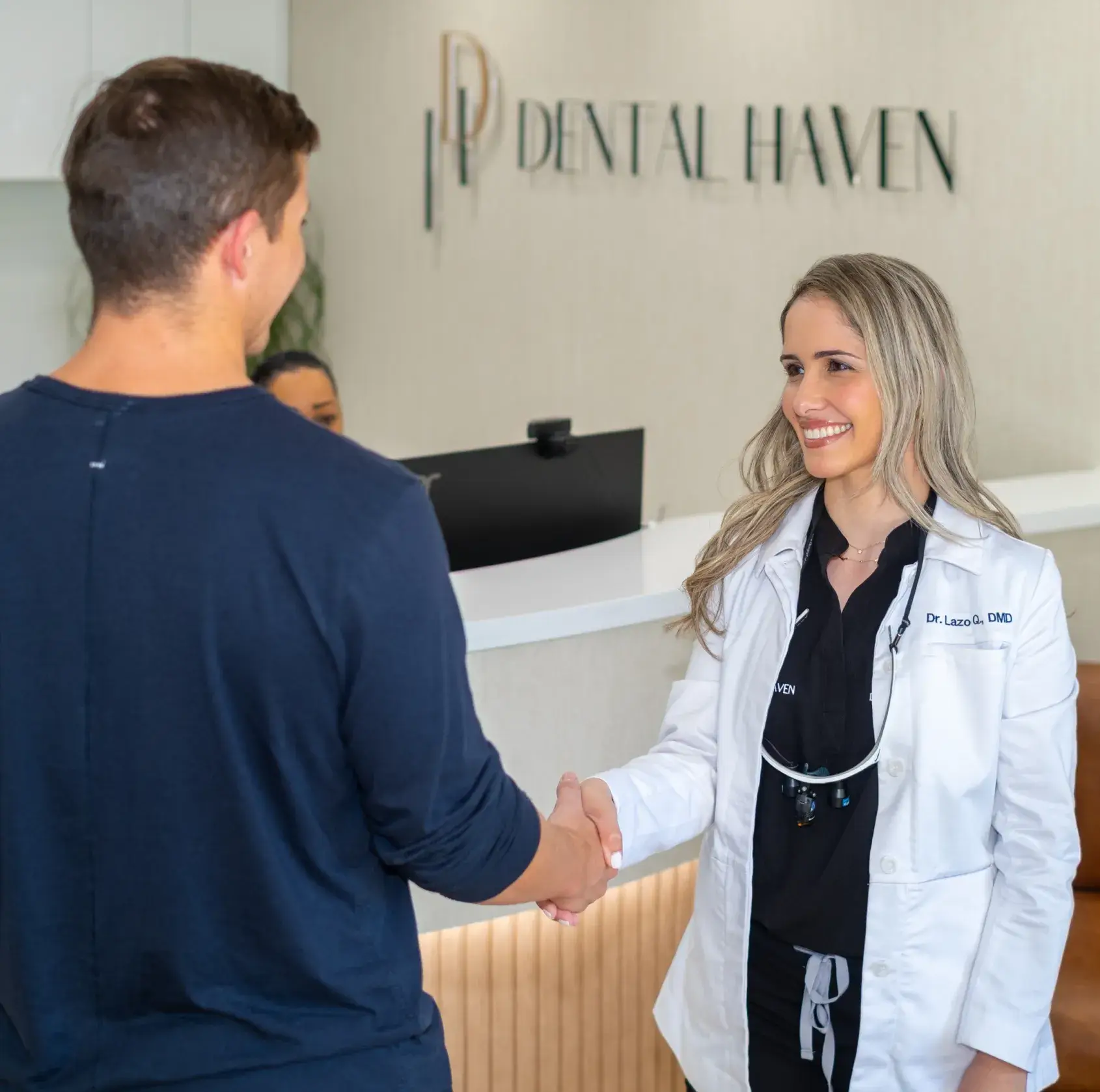 A dentist in a white coat smiles and shakes hands with a patient at a dental office.