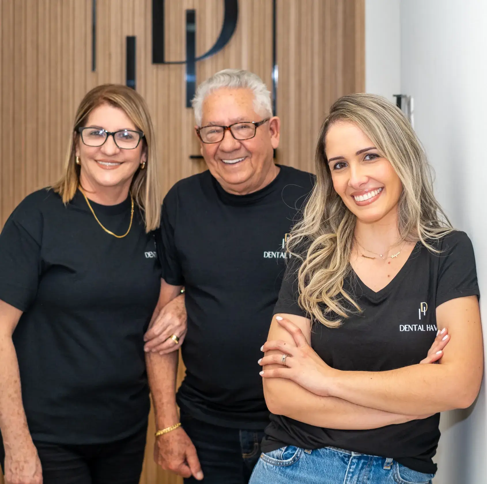 Three people wearing matching black shirts smile at the camera inside a dental office.