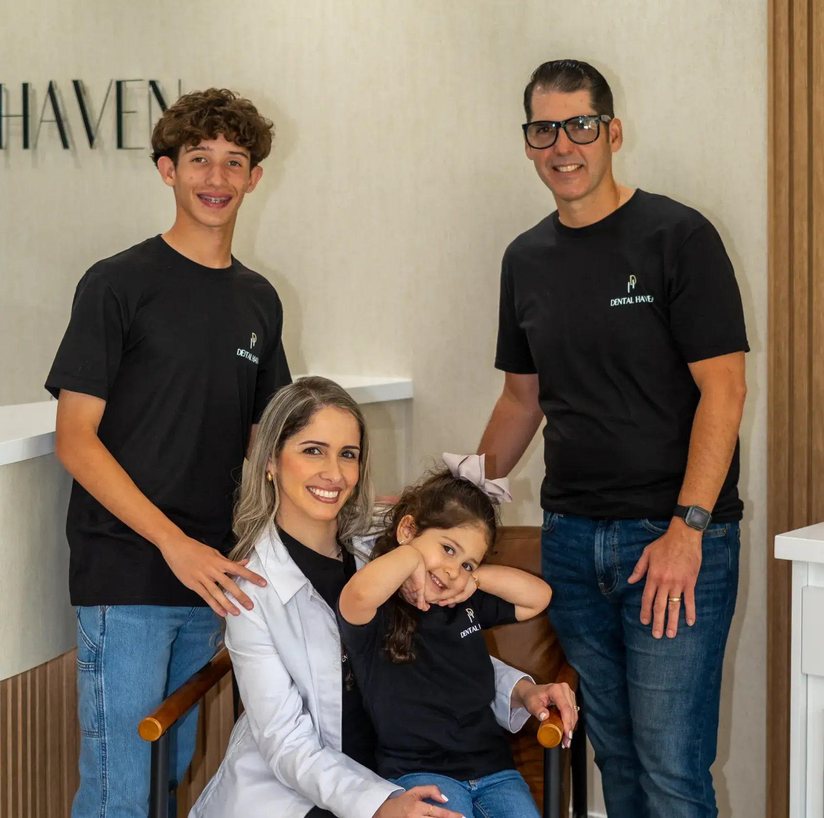 A smiling family of four poses together inside a room, with a young girl sitting on a chair.