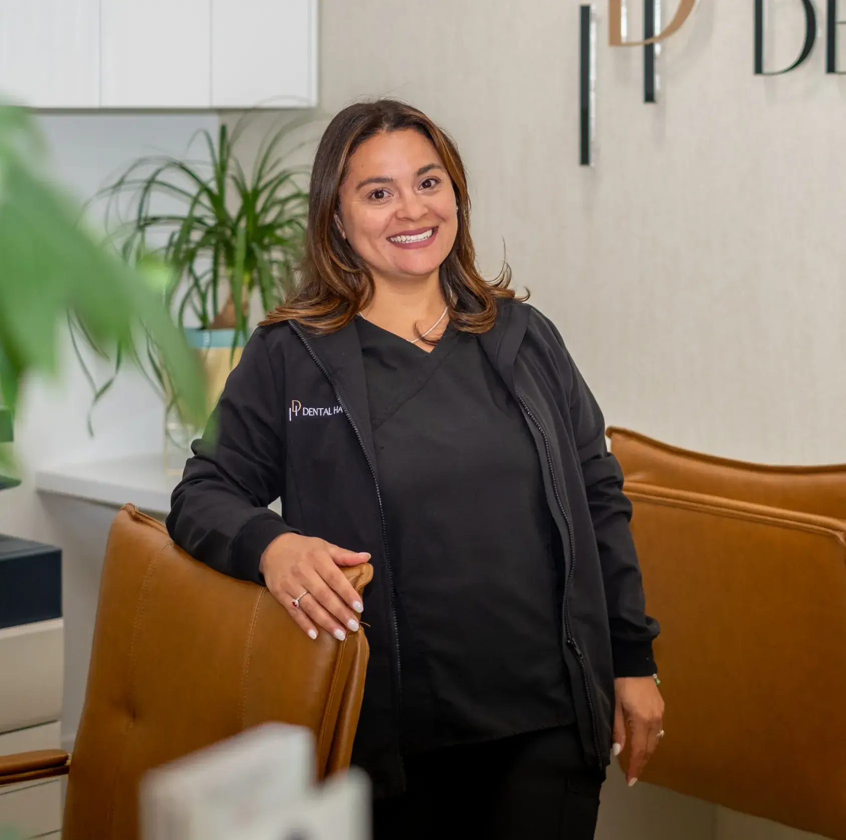 A woman in a black jacket smiles while standing beside a brown chair in an office.