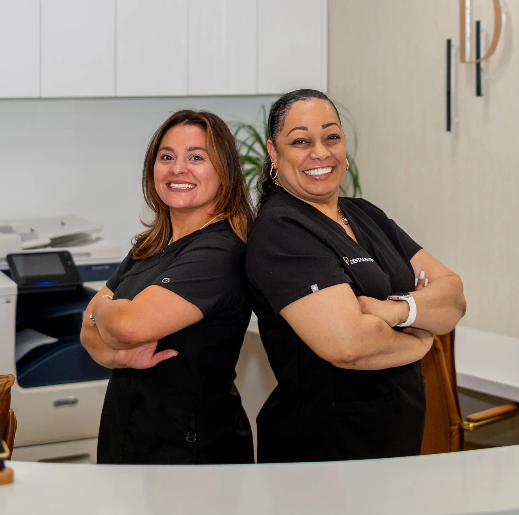 Two smiling women in black uniforms stand back-to-back with arms crossed in an office setting.