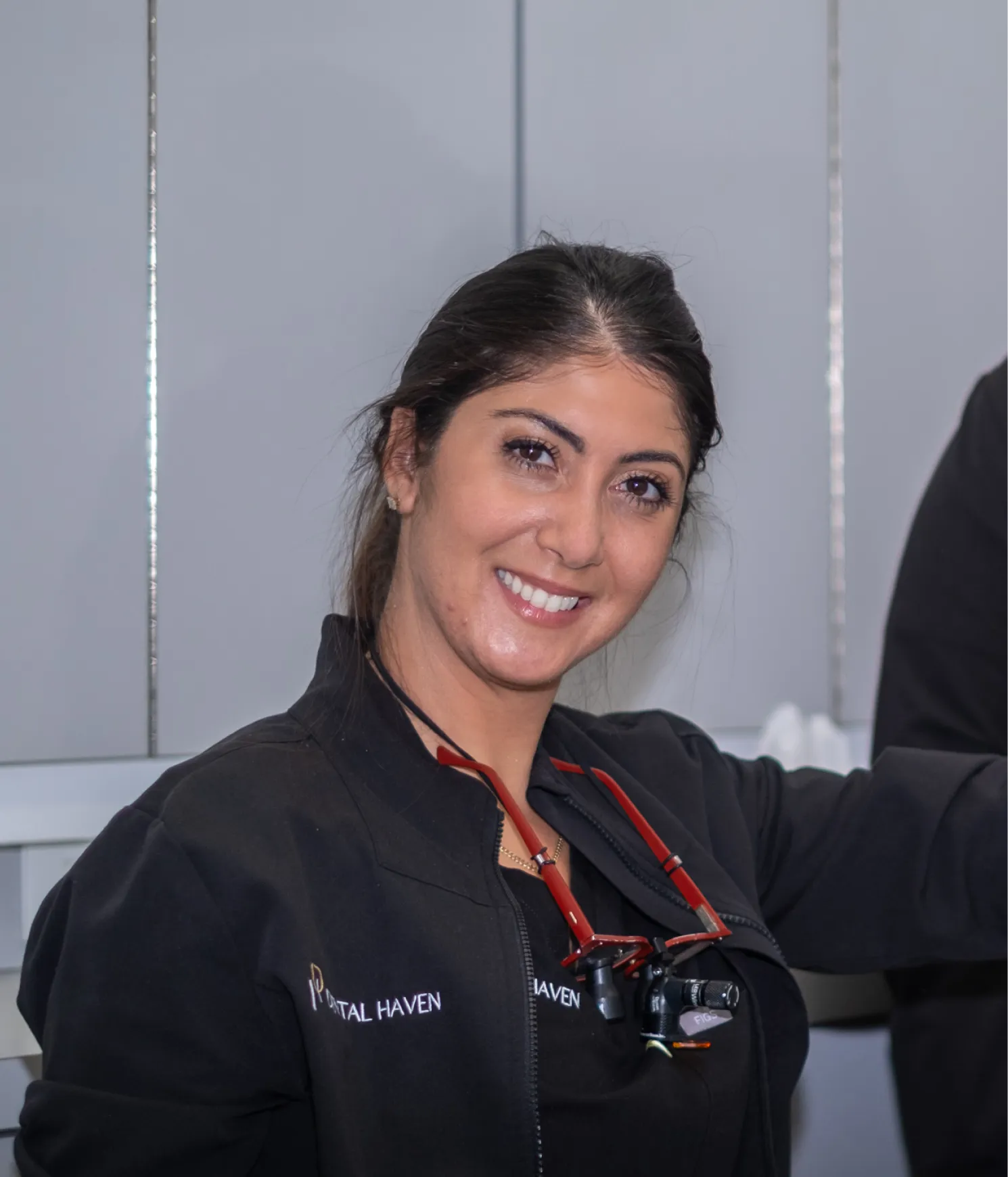 Smiling woman in black dental uniform with magnifying glasses around her neck stands in a dental office.