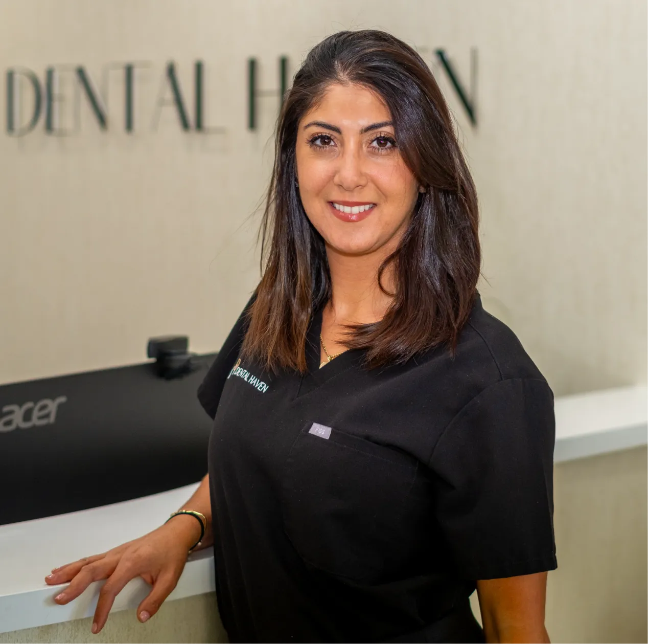 Smiling woman in black uniform stands by a reception desk at a dental office.