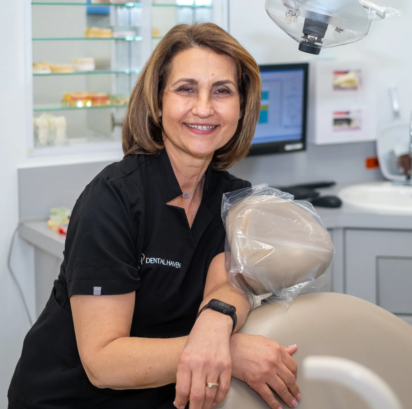 A smiling dental professional stands beside a dental chair in an examination room.