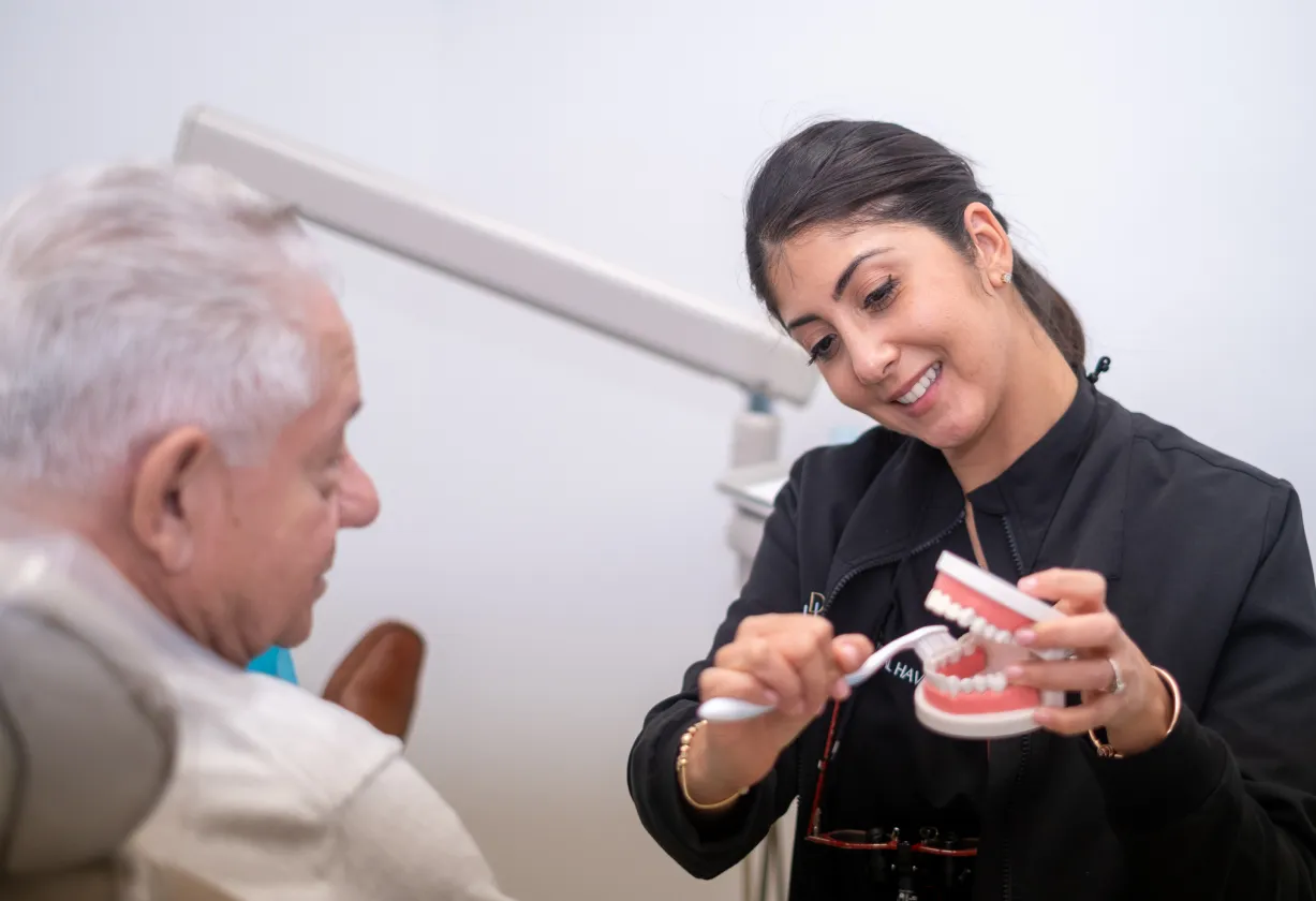 A dental professional demonstrates brushing techniques to an older man using a dental model.