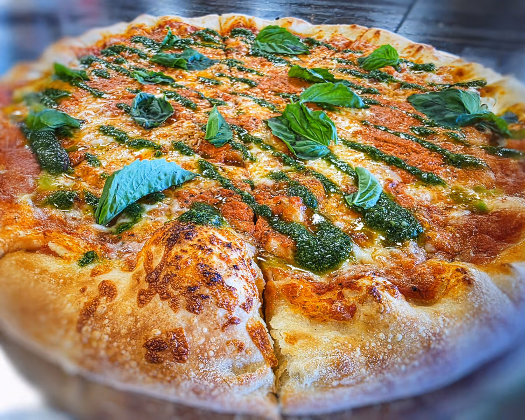 Close-up of a pizza with melted cheese, tomato sauce, fresh basil leaves, and drizzled green pesto sauce on a wooden table.
