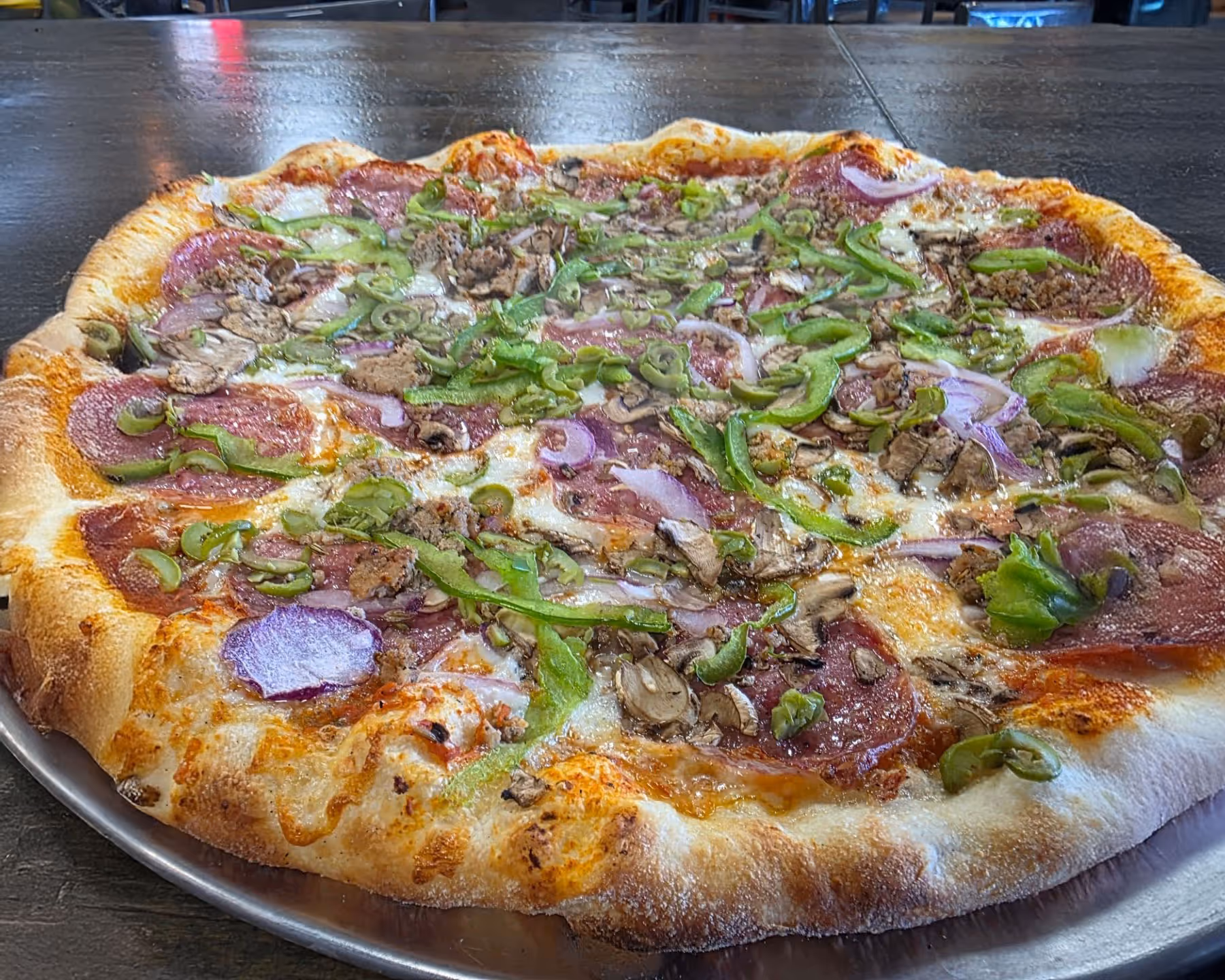 Fresh pizza topped with pepperoni, green bell peppers, mushrooms, red onions, and sausage on a metal tray.