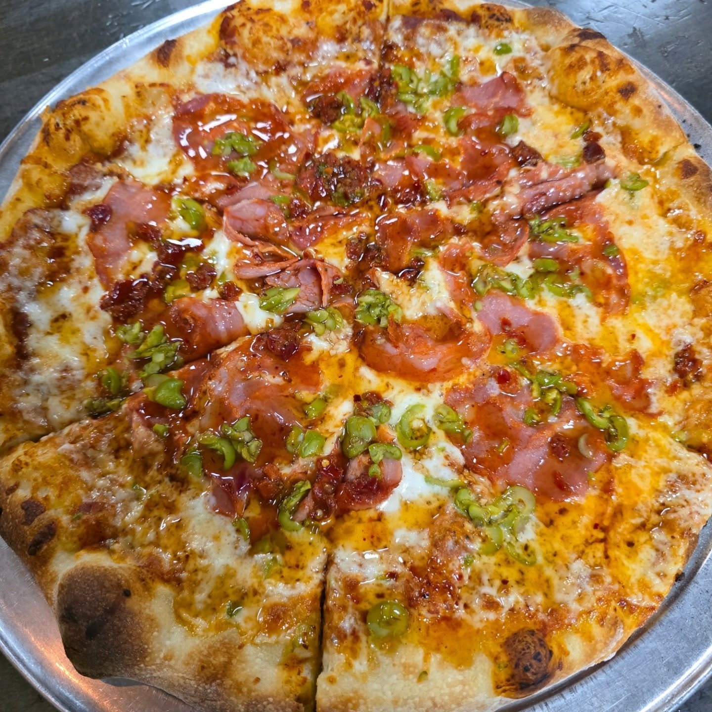 Whole pizza topped with cheese, slices of pepperoni, ham, chopped green peppers, and red chili flakes on a metal tray.