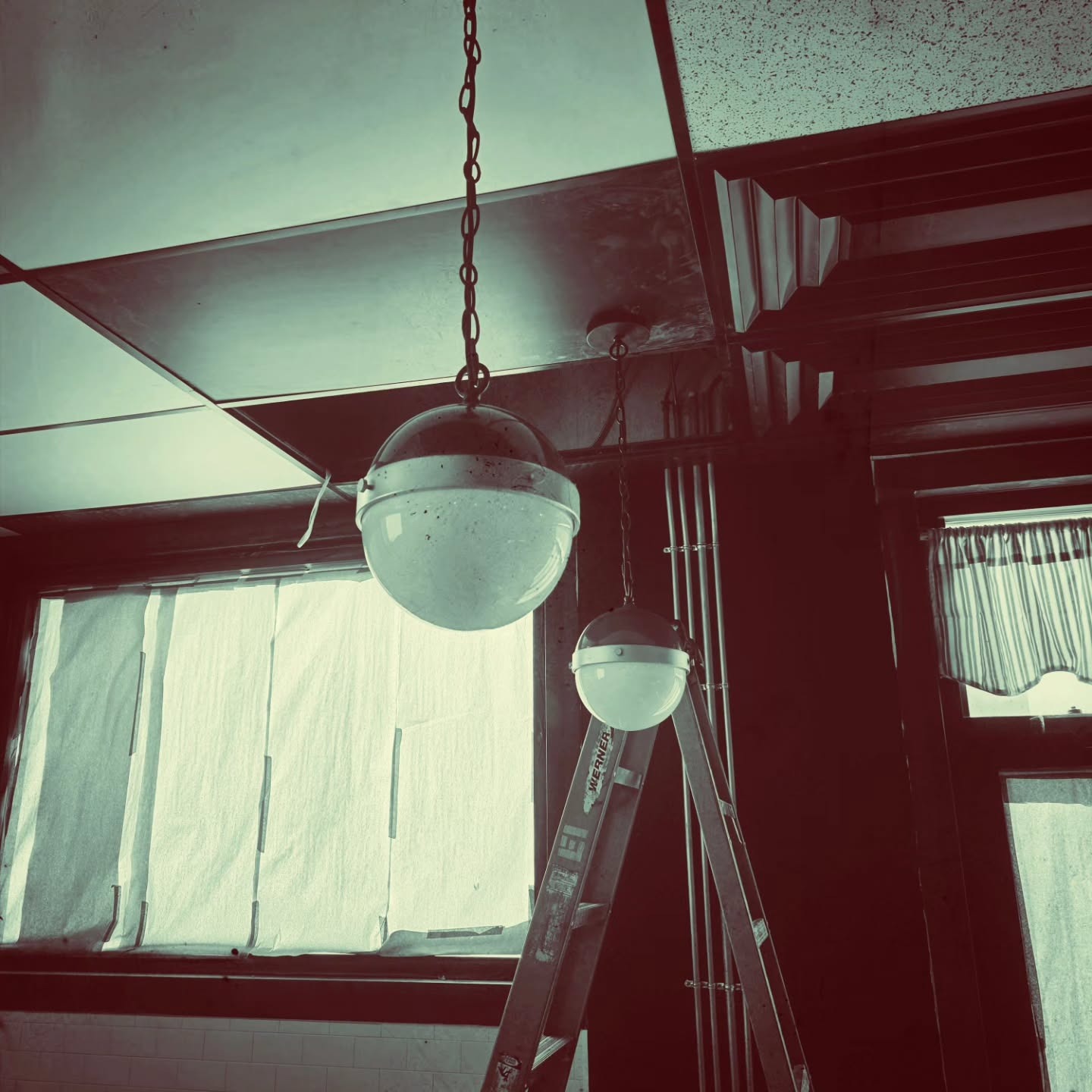 Two round pendant lights hanging from a ceiling near a window covered with paper and a metal ladder positioned underneath.