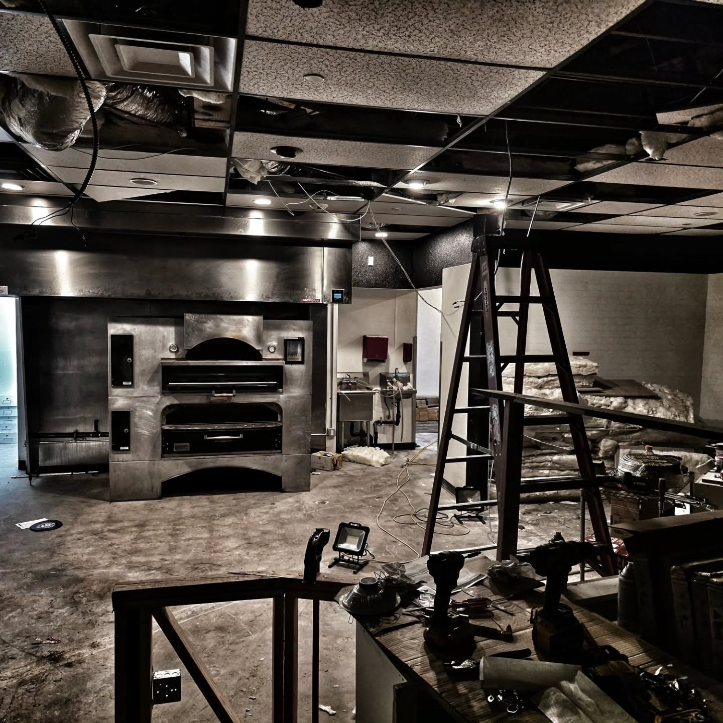 Industrial kitchen under construction with a large stainless steel oven, a red ladder, exposed ceiling, and tools on a workbench.