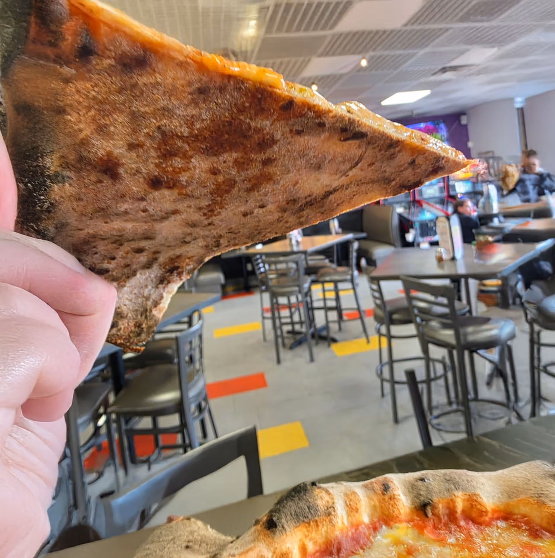Hand holding up a slice of pizza showing the charred underside crust in a pizzeria with tables and chairs in the background.