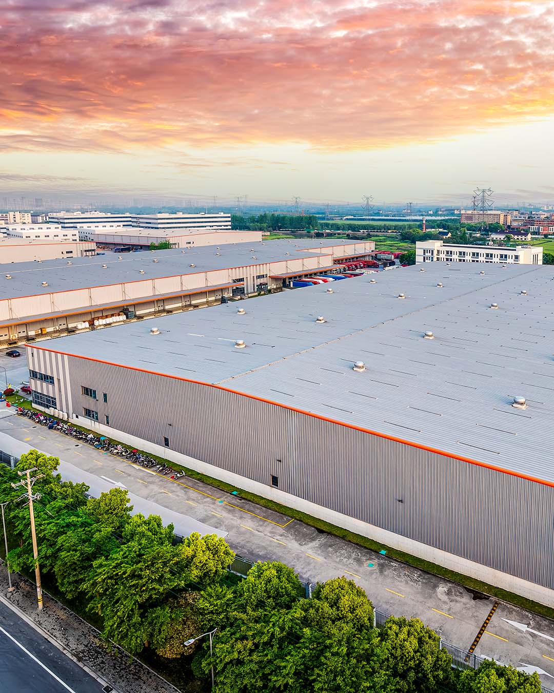 Aerial view of large industrial warehouses with a road, parked bikes, and green trees under a colorful sunset sky.