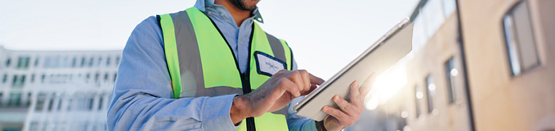 Construction worker in neon safety vest using a tablet outside near buildings.
