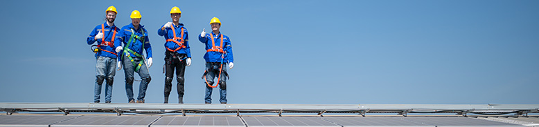4 roofing professionals wearing saftey gear and waving to the camera from a commercial rooftop.