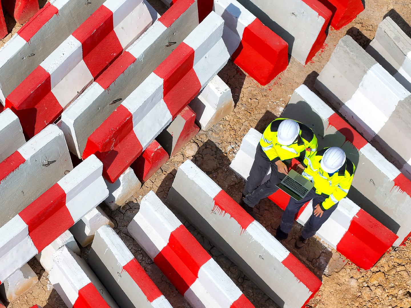 Two construction workers in yellow jackets and helmets sit on concrete barriers while looking at a laptop.