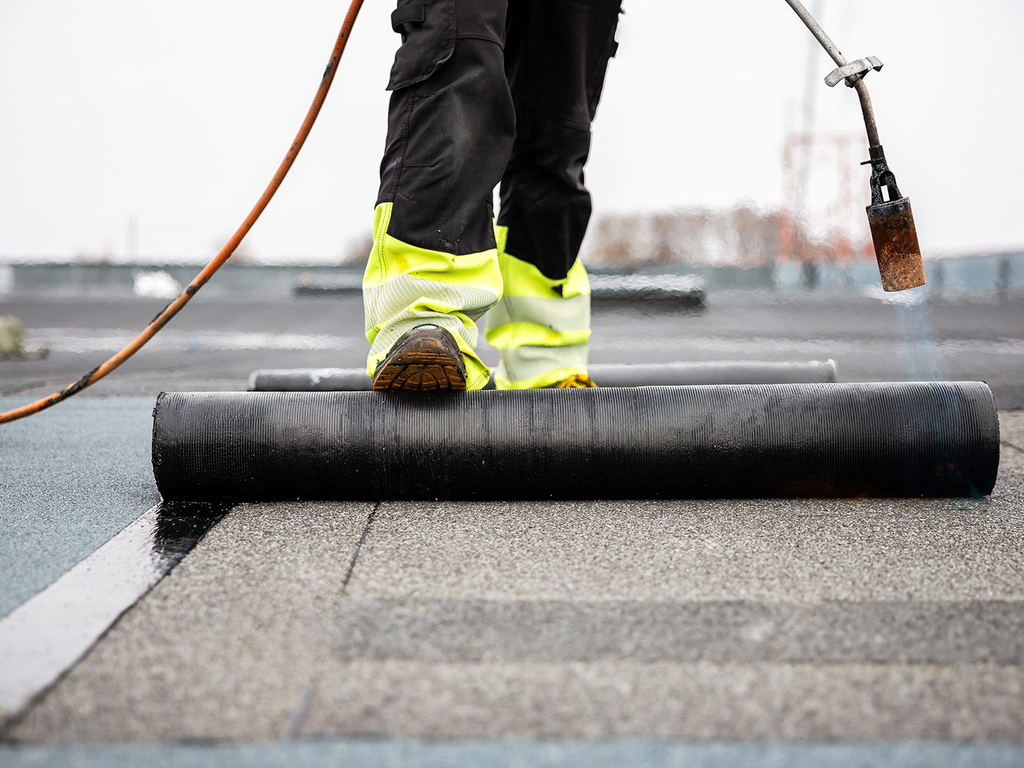 Worker wearing yellow reflective pants rolling out black roofing material on flat roof with torch.