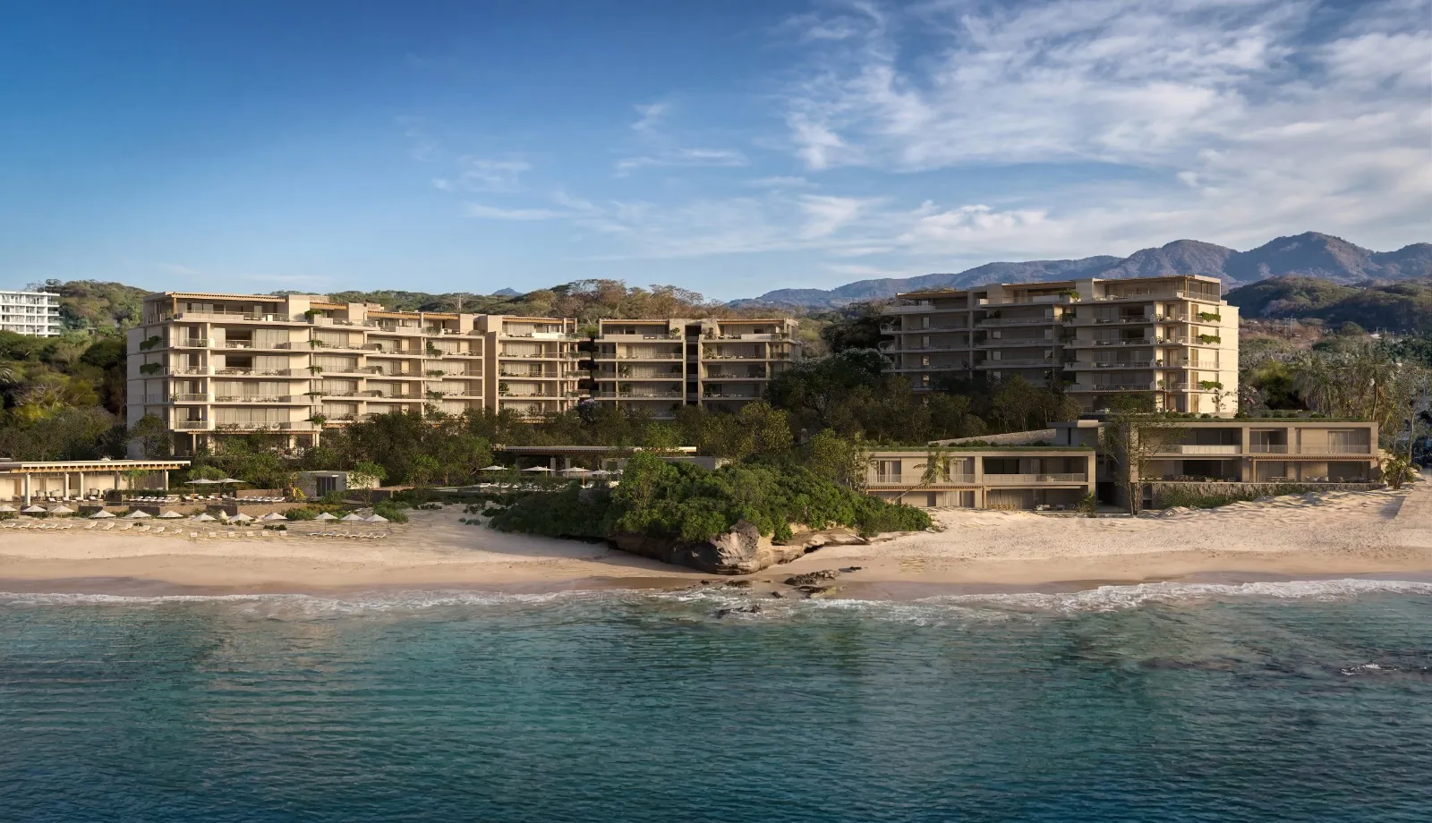 Modern beachfront resort with multiple low-rise buildings, sandy beach, blue ocean, and forested hills in the background under a clear sky.