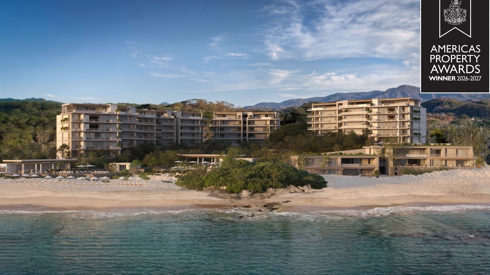 Modern beachfront resort with multiple low-rise buildings, sandy beach, blue ocean, and forested hills in the background under a clear sky.