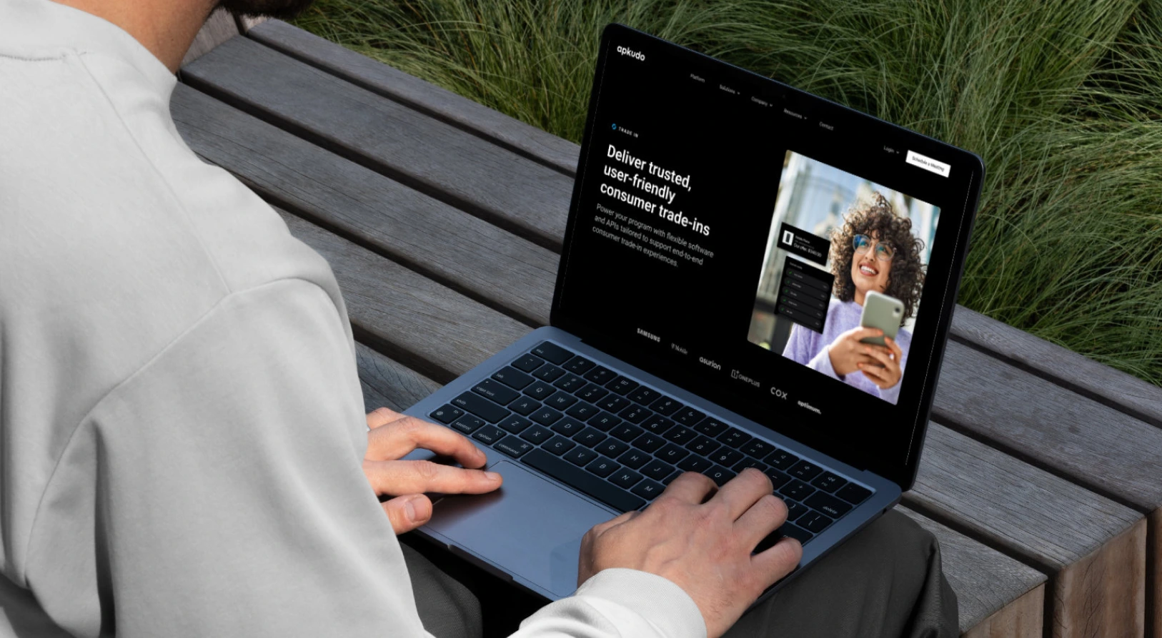 Person using a laptop on an outdoor wooden bench, viewing a webpage about trusted user-friendly consumer trade-ins with an image of a smiling woman holding a smartphone.