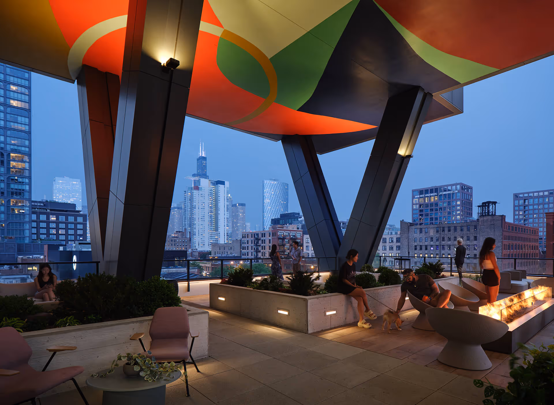 Rooftop terrace at dusk with modern art ceiling, city skyline in the background, people socializing, and a long fire pit.