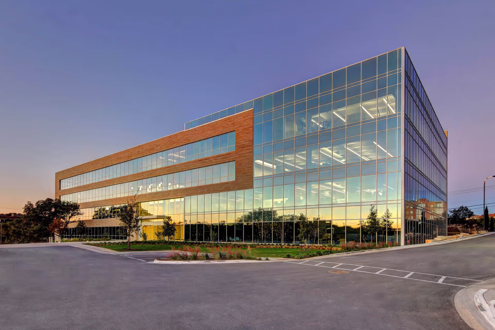 Modern glass office building with wood accents reflecting sunset, surrounded by a landscaped parking area.