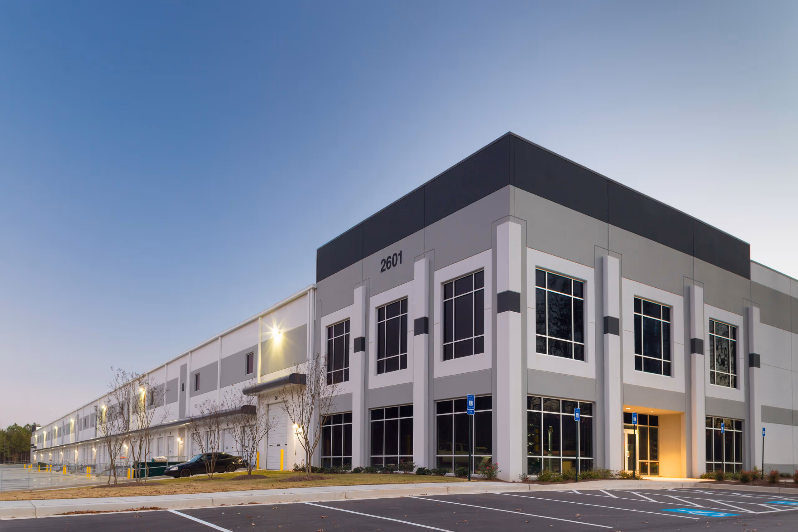 Modern industrial distribution center building with large windows, loading dock doors, and parking lot marked for handicapped spaces under early evening sky.