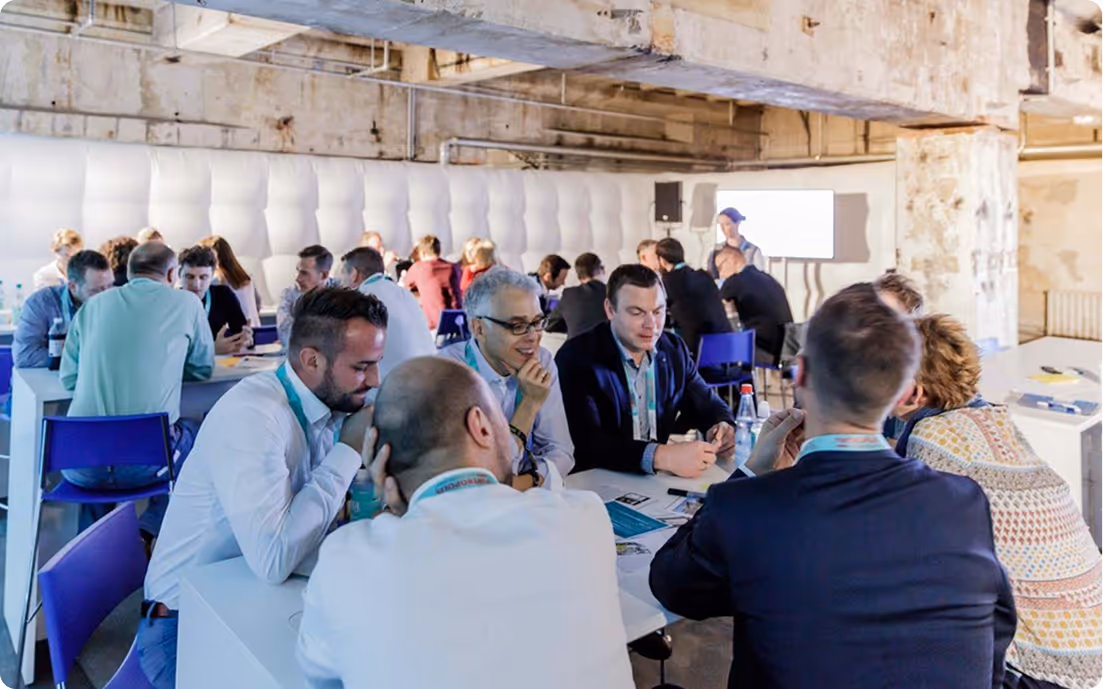 Mehrere Gruppen von Menschen sitzen in einem hellen Raum an Tischen und führen Diskussionen bei einem Workshop oder Meeting.