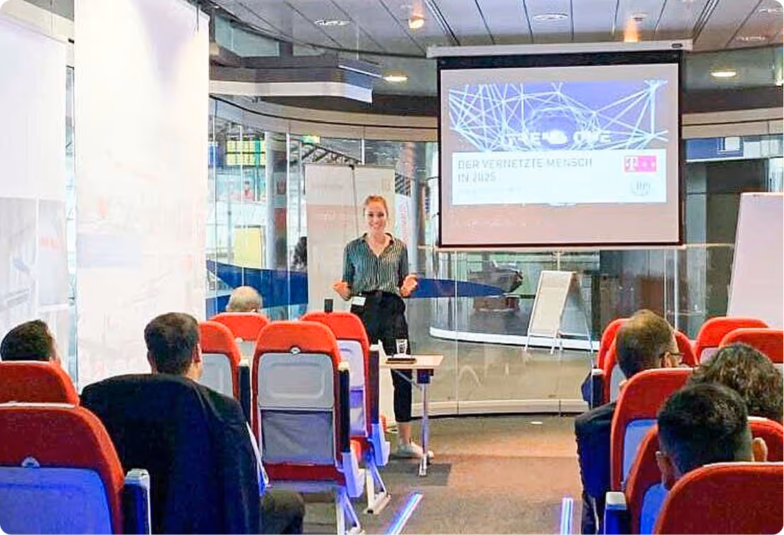 Frau hält eine Präsentation vor einem Publikum in einem modernen Raum mit roten Sitzen und einem Projektorbildschirm.