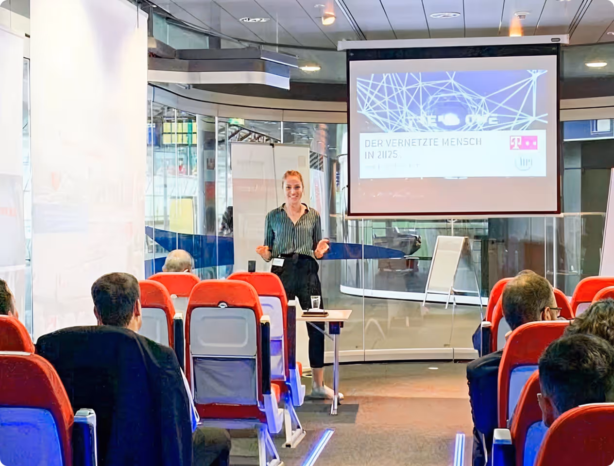 Frau hält Präsentation vor sitzendem Publikum mit rotem Sitzstuhl im modernen Konferenzraum, auf Leinwand steht 'Der vernetzte Mensch in 2125'.