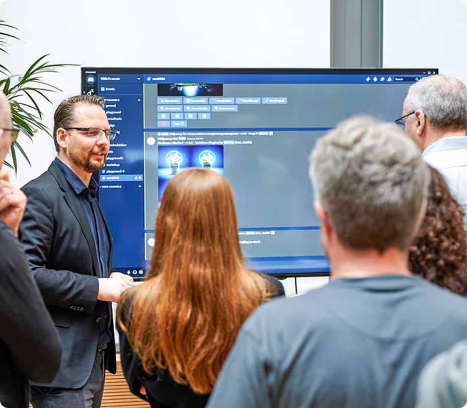 Eine Gruppe von Menschen schaut auf einen großen Bildschirm, auf dem eine Präsentation mit Symbolen von Glühbirnen sichtbar ist.
