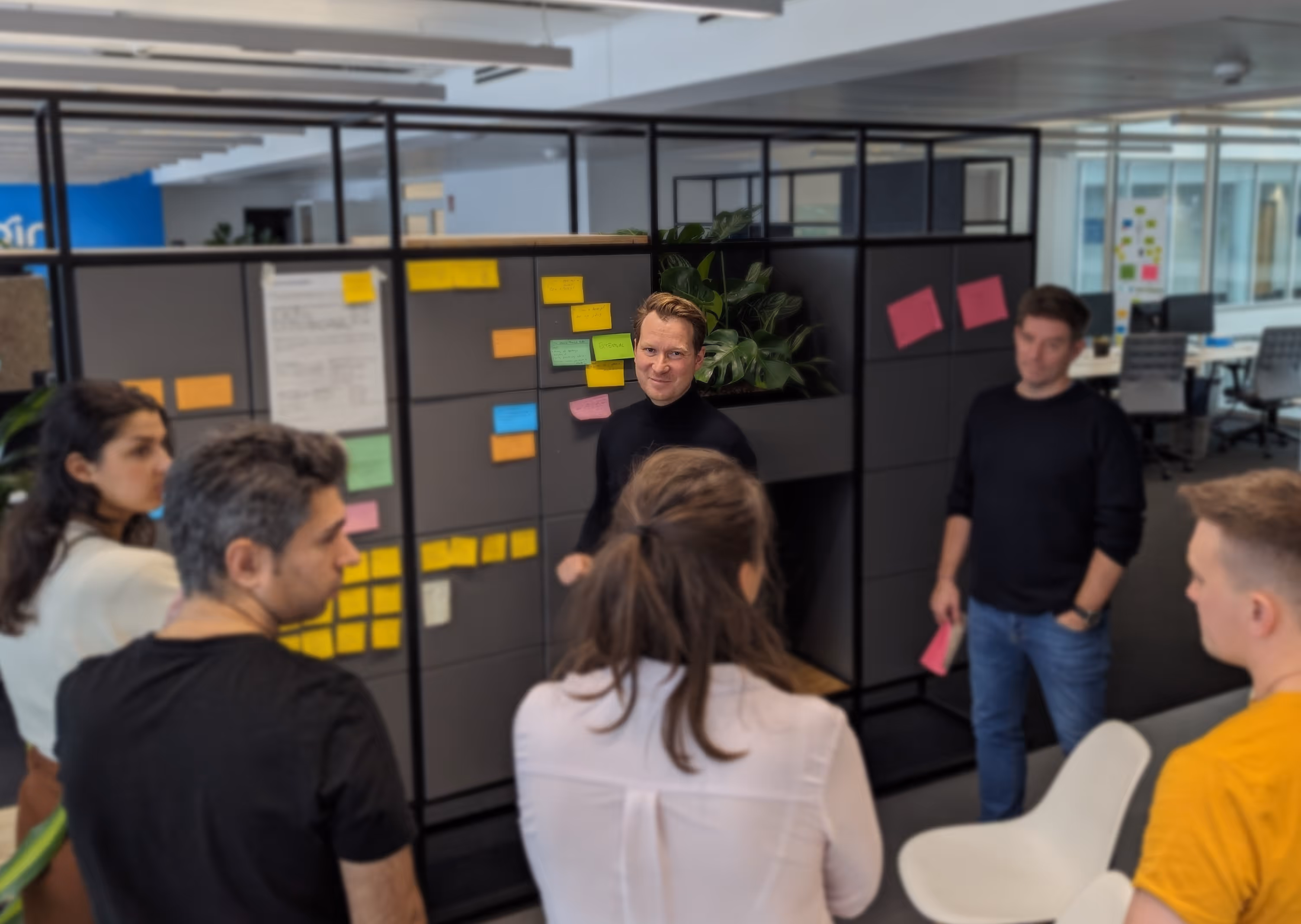 Team von sechs Personen diskutiert vor einer Wand mit bunten Haftnotizen in einem modernen Büro.
