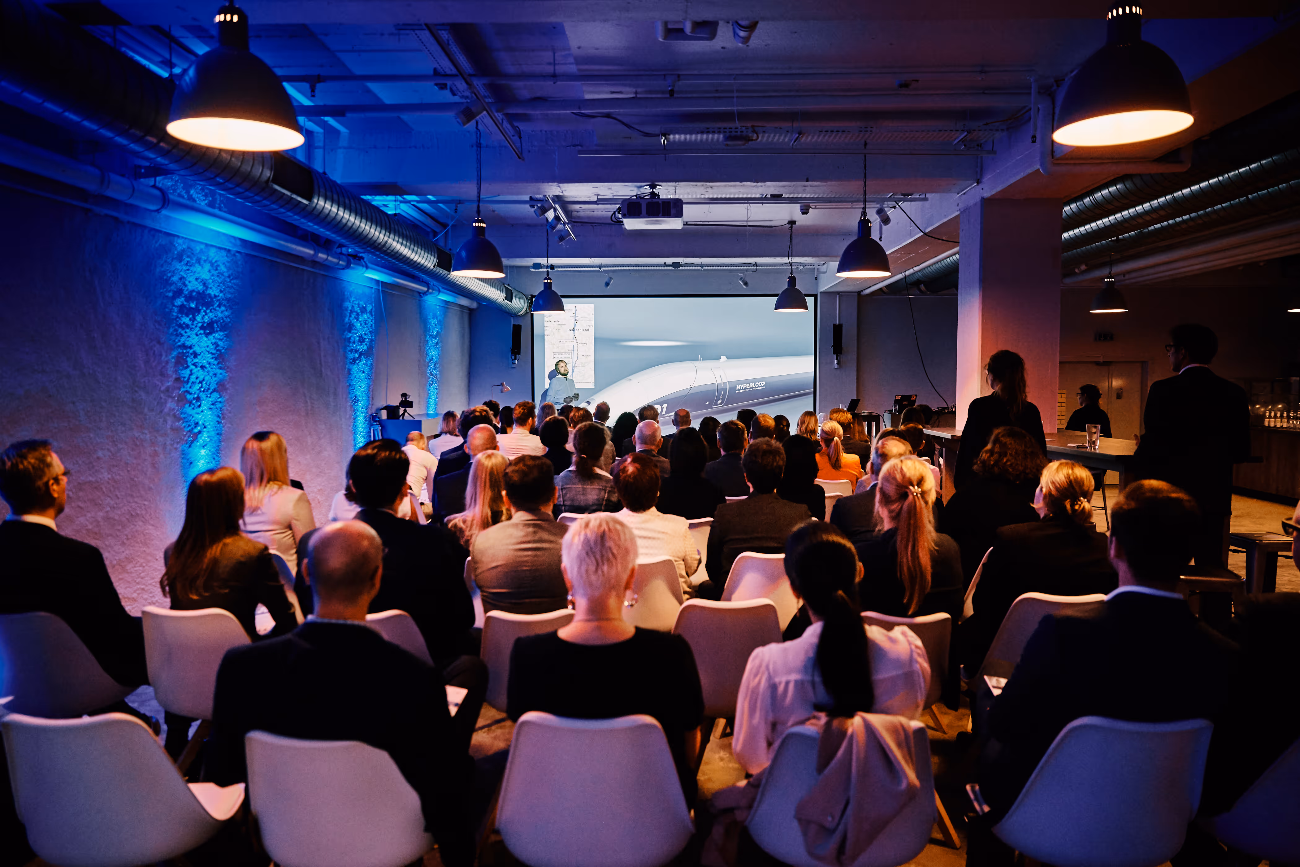 Zuhörer in einem Konferenzraum sehen eine Präsentation über Hyperloop-Technologie.