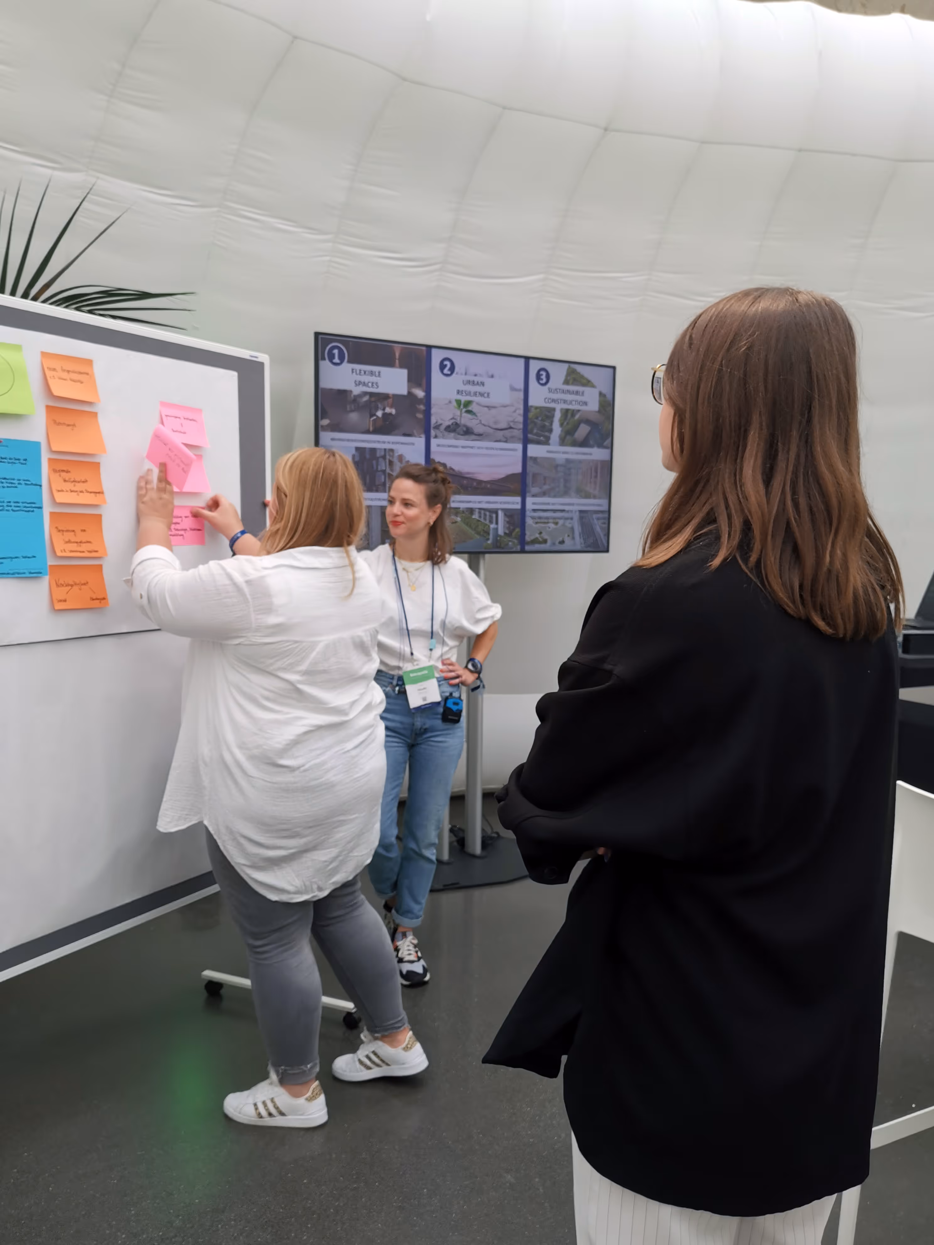 Drei Frauen diskutieren vor einem Whiteboard mit farbigen Haftnotizen und einem Bildschirm mit Bauplänen.
