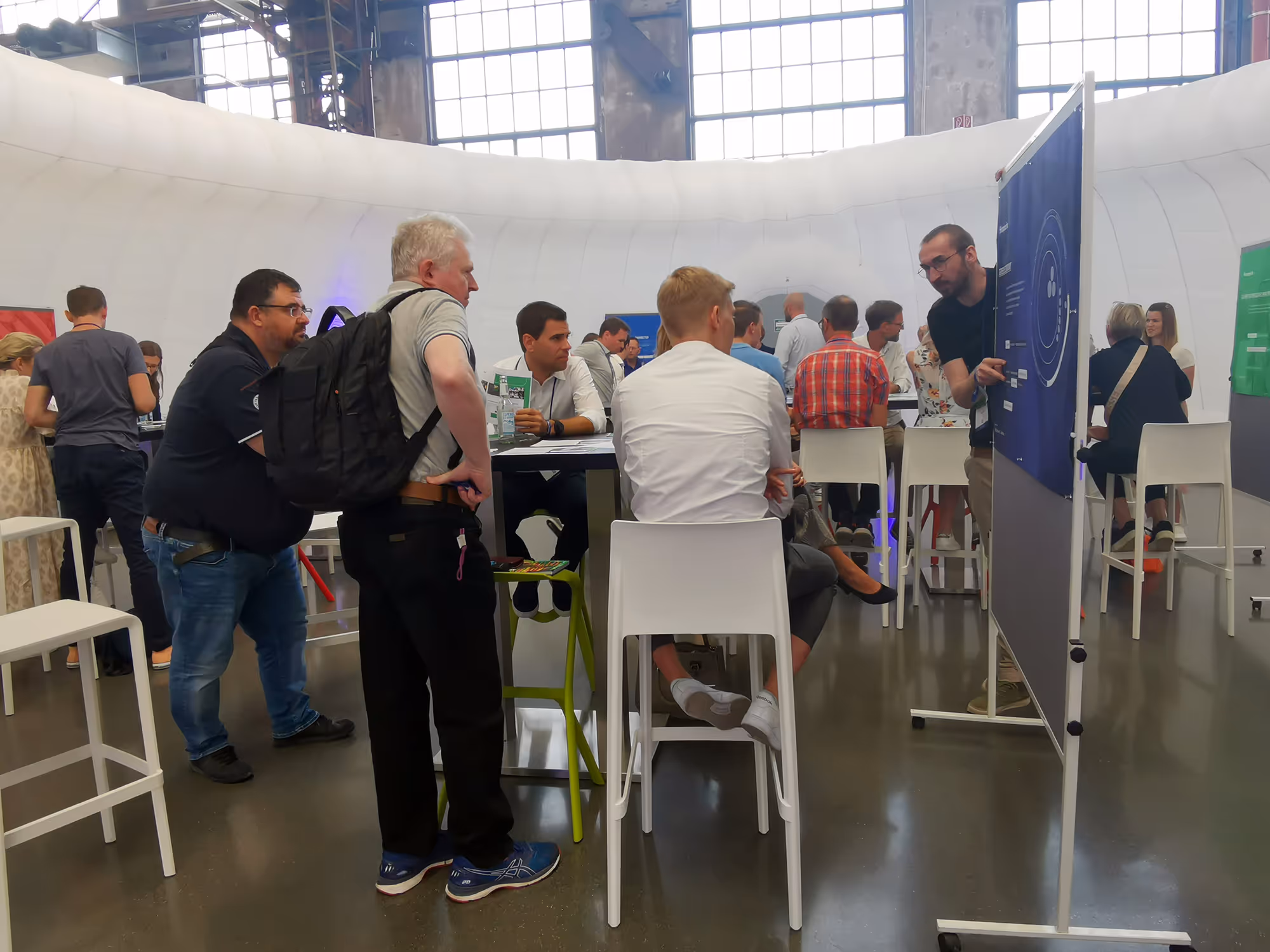 Menschen diskutieren an hohen Tischen in einem modernen, hellen Veranstaltungsraum mit großen Fenstern.