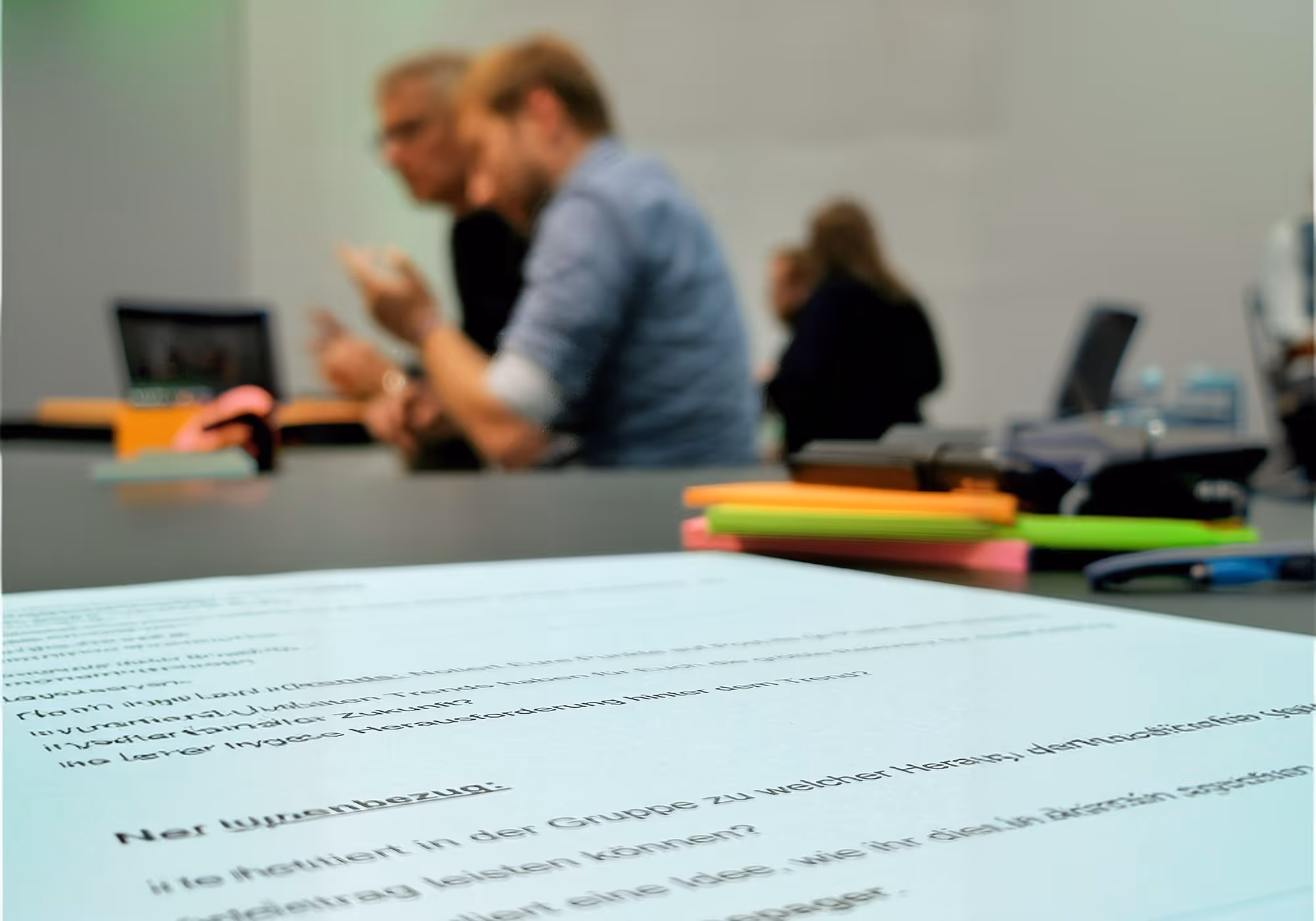 Vordergrund mit deutschem Textdokument, im Hintergrund unscharfe Leute bei Besprechung am Tisch.