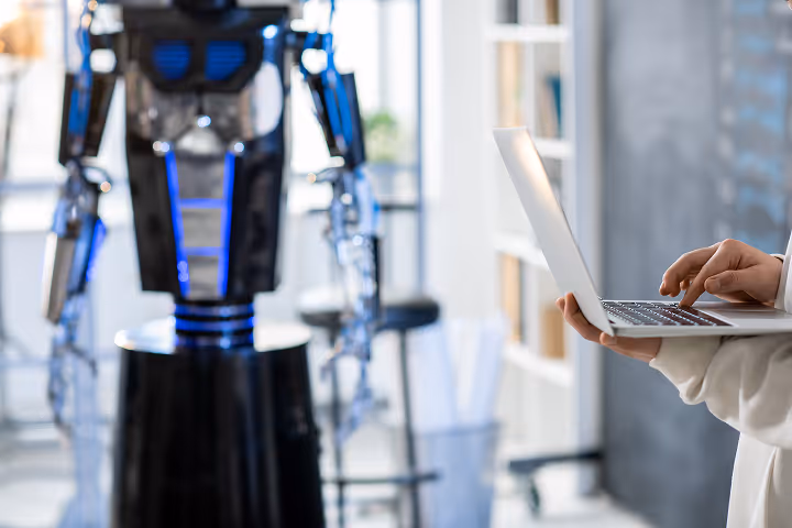 Eine Person bedient Laptop nahe einem stehenden humanoiden Roboter mit blauen Details.