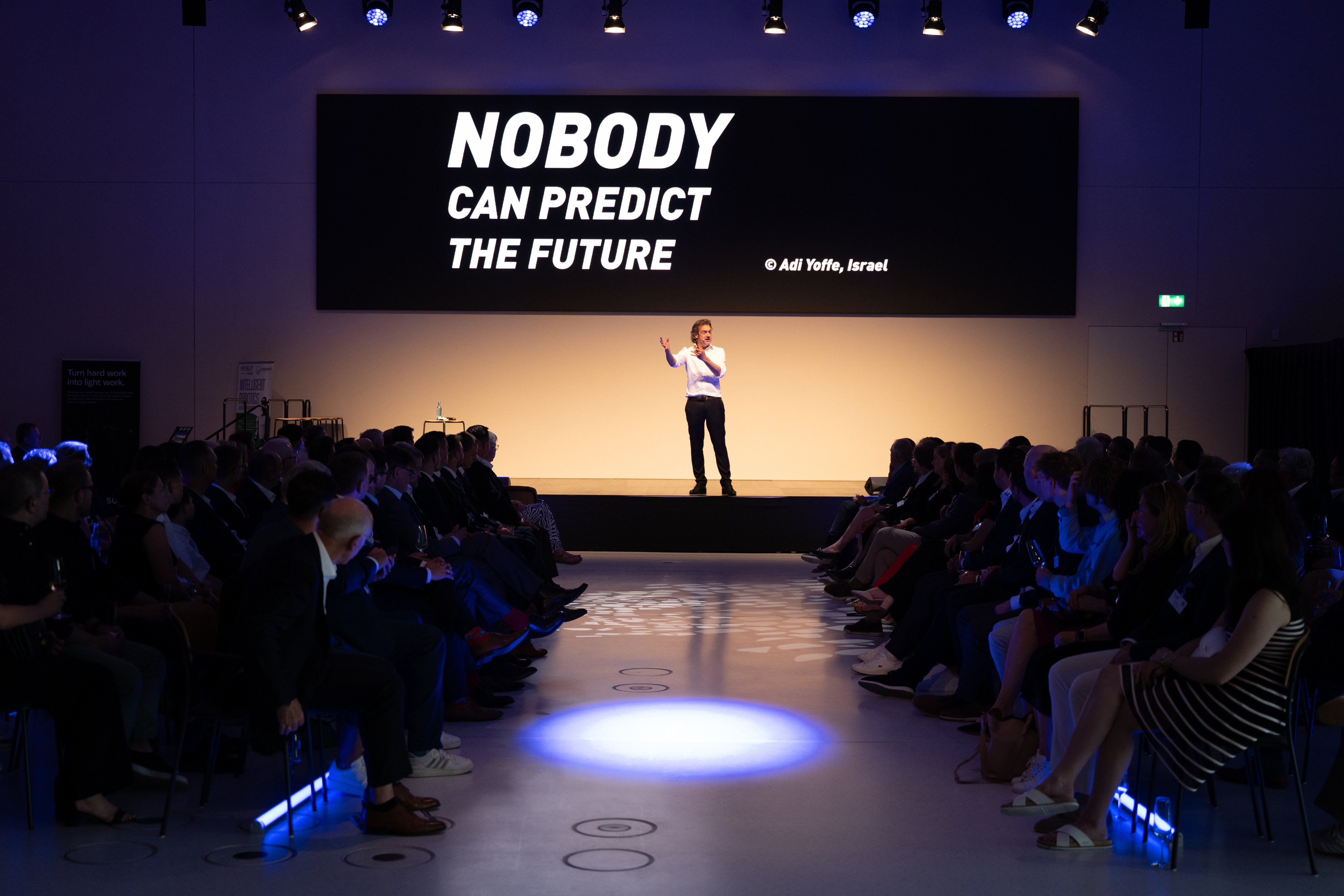 Ein Redner auf einer Bühne vor Publikum vor einer Leinwand mit Text Nobody can predict the future.
