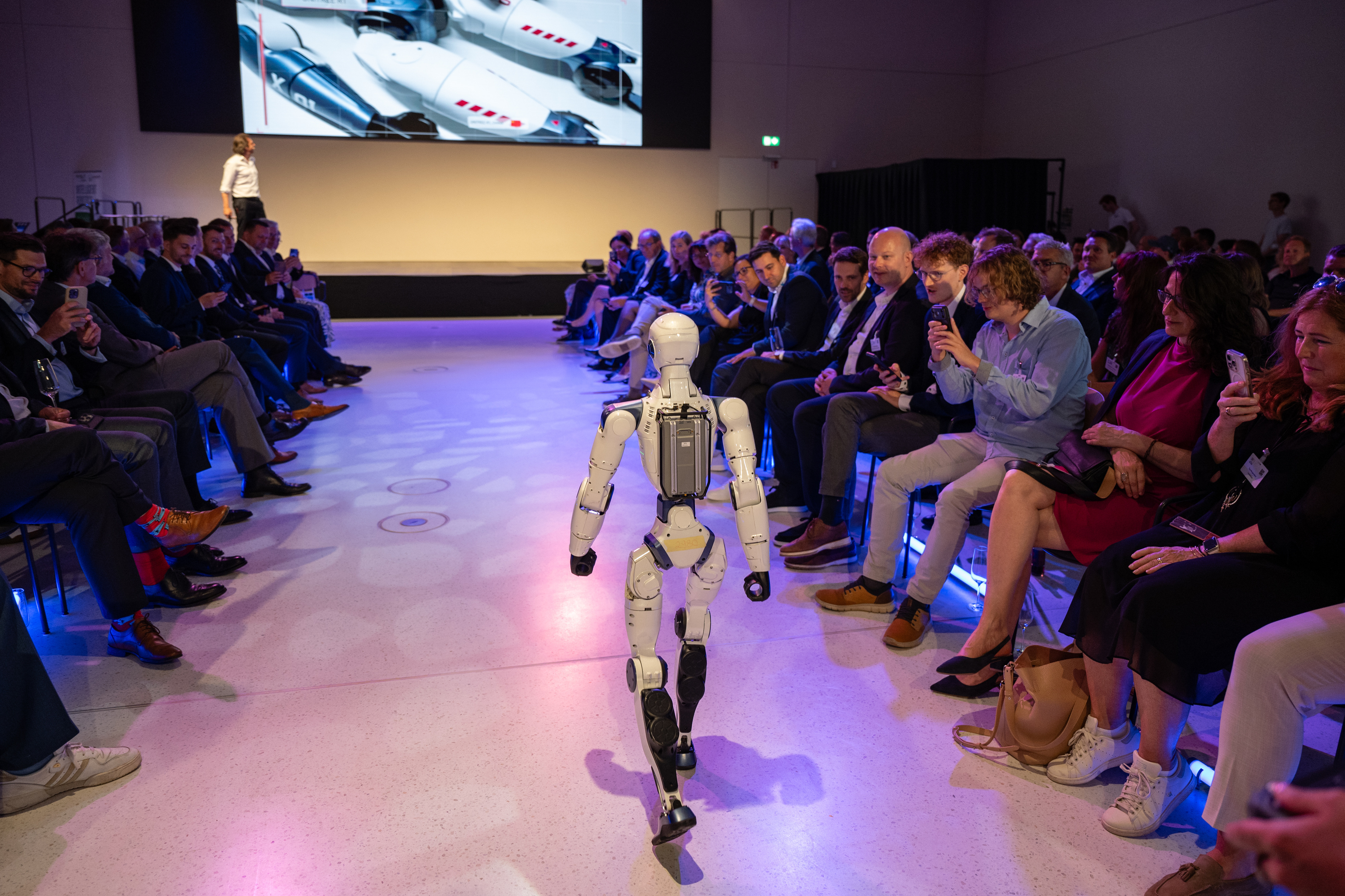 Ein humanoider Roboter läuft auf einer Bühne zwischen Publikum, das Fotos macht und zuschaut.