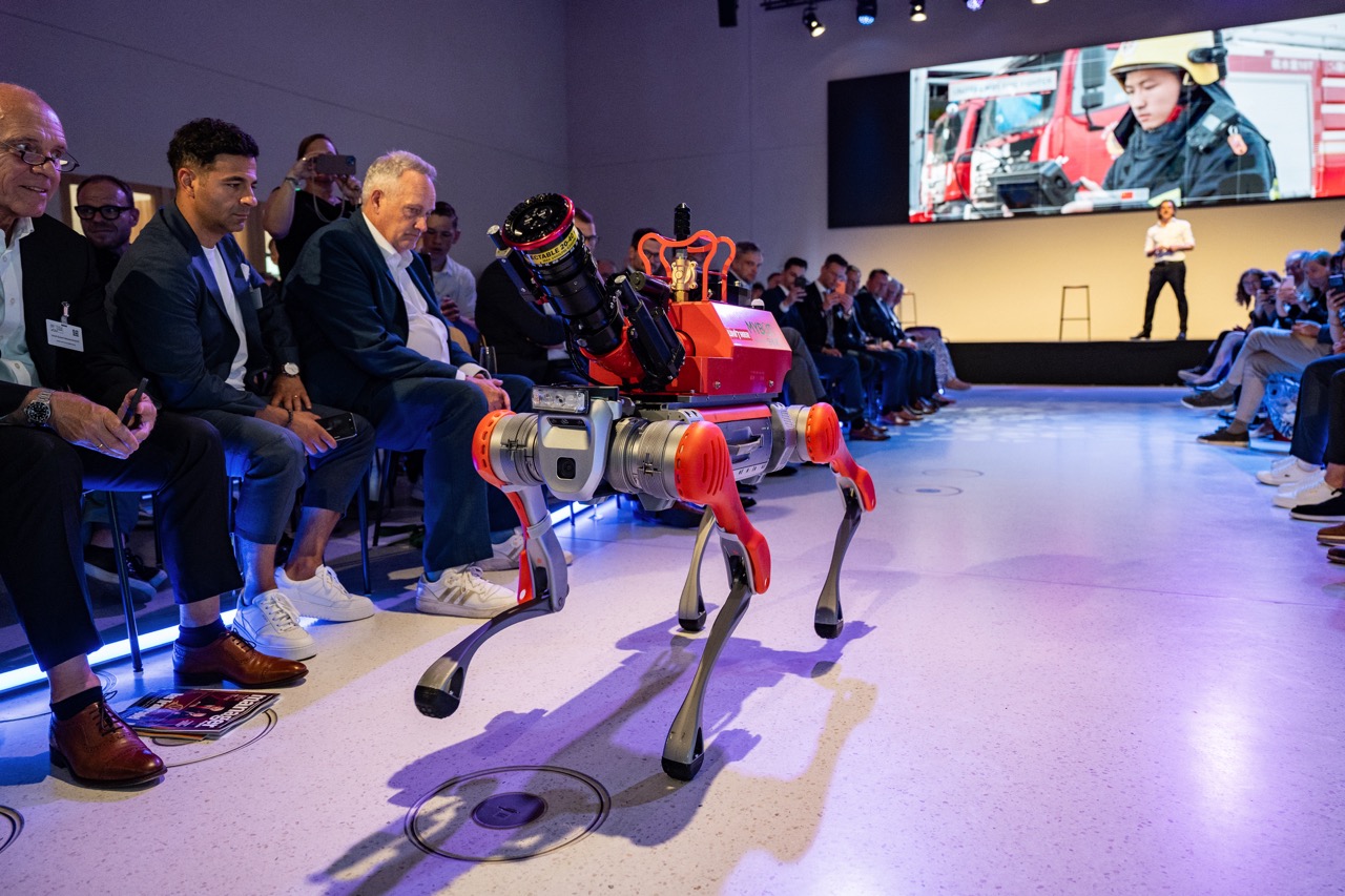 Vierbeiniger Roboter in Rot und Weiß läuft durch Raum mit Publikum bei Präsentation.