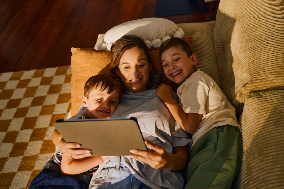 A woman lying on a couch with two young boys, all smiling and looking at a tablet she is holding.