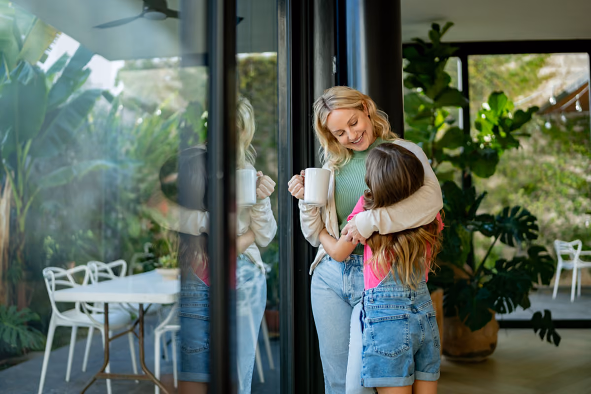 Woman holding a mug smiling and hugging a girl wearing a pink shirt and denim overalls indoors.