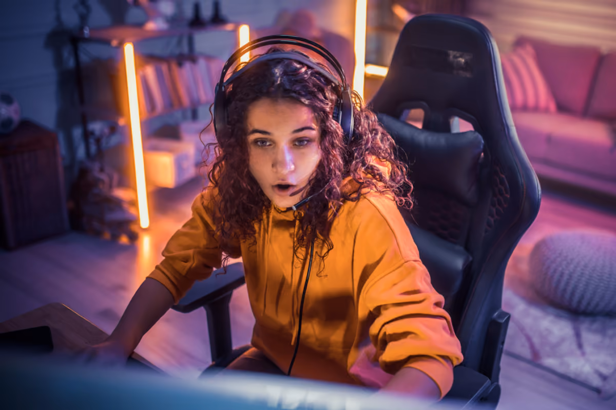 Young woman with curly hair wearing a gaming headset and orange hoodie intensely focused on a computer screen while sitting in a gaming chair in a dimly lit room with neon lights.