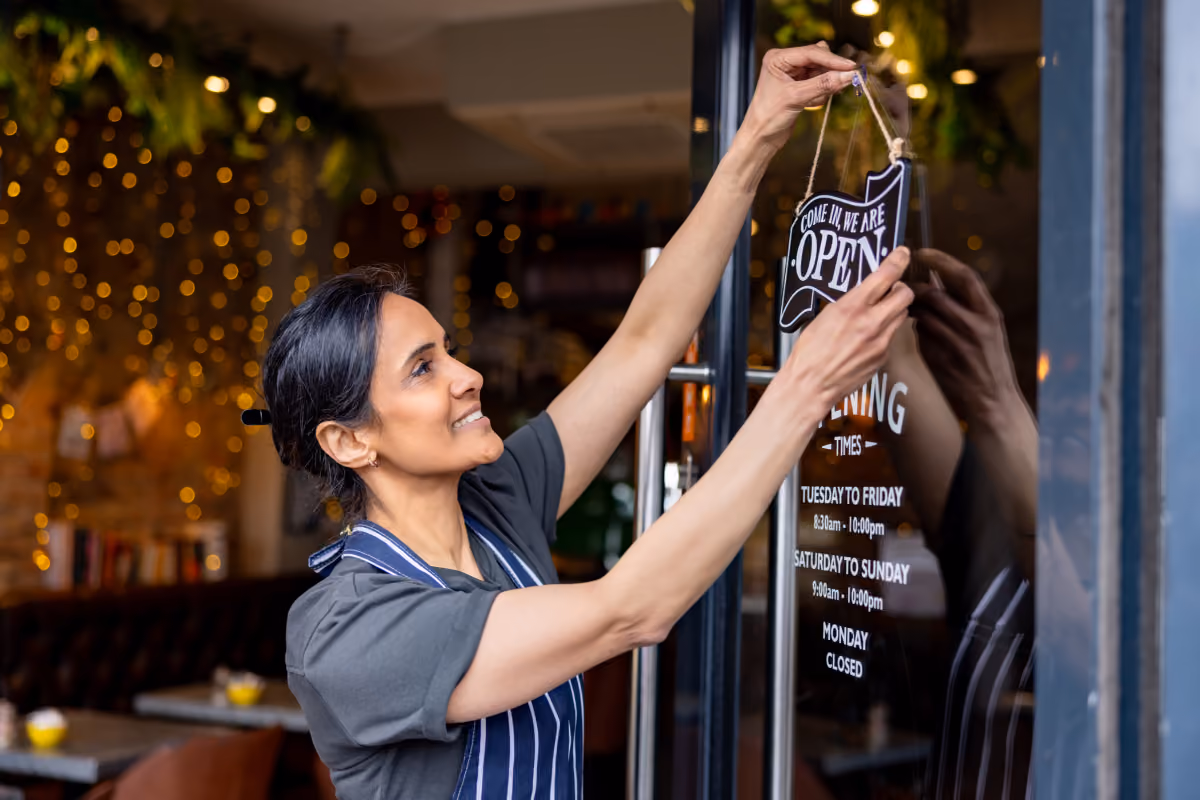 A woman with dark hair in a striped apron hangs an 'Open' sign on a glass door displaying business hours.