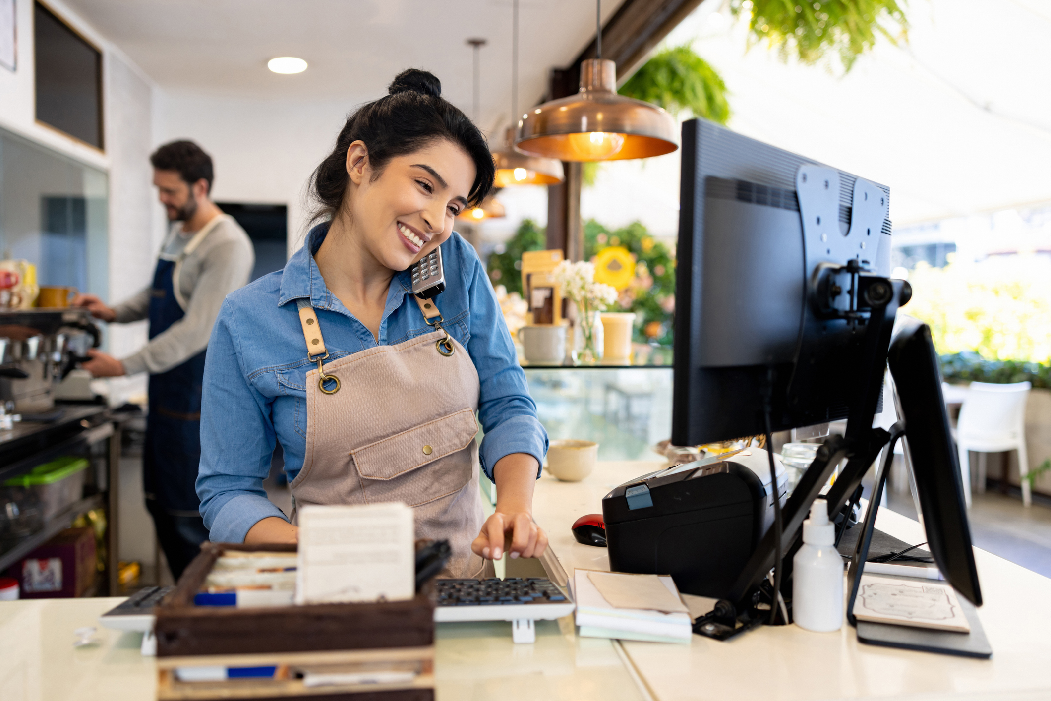 A smiling female barista with a beige apron and blue shirt is multitasking by holding a phone between her shoulder and ear while typing on a keyboard at a coffee shop counter with a computer and receipt printer nearby.
