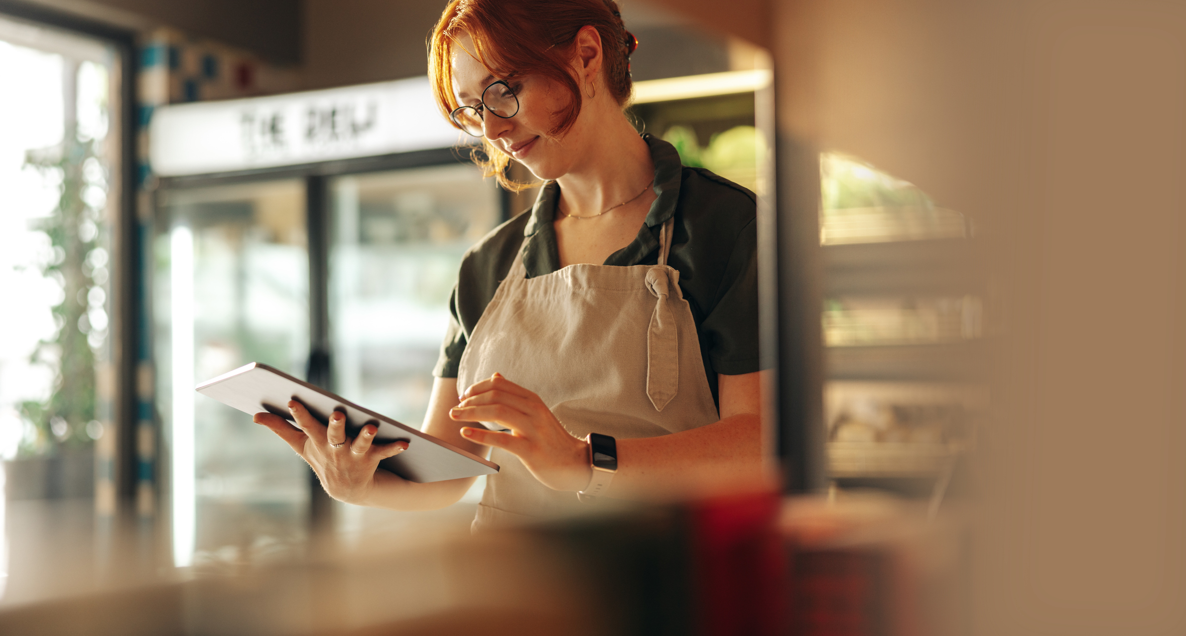 Young woman with red hair wearing glasses and an apron using a digital tablet in a bright café or shop.