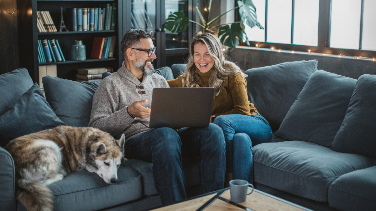A middle-aged man and woman sitting on a gray couch looking at a laptop and smiling, with a large dog resting beside them and a bookshelf in the background.
