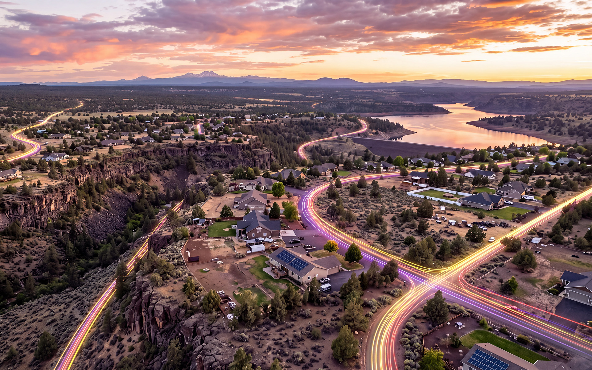 Aerial view of a suburban area at sunset with winding roads showing colorful light trails representing fiber internet connectivity, a river or lake surrounded by rocky terrain and trees, residential houses, and distant mountains under a partly cloudy sky.